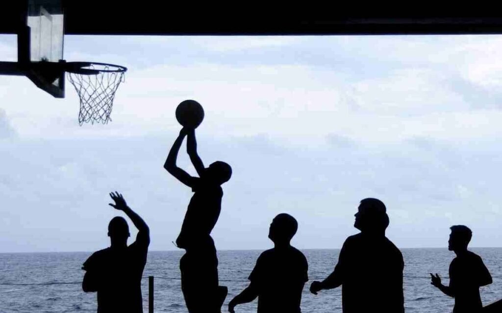 Five silhouettes of people playing basketball, with the sea in the distance.