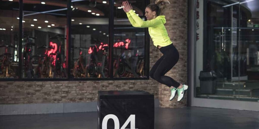 Woman in a gym, jumping up into a high box, demonstrating multiple components of fitness.